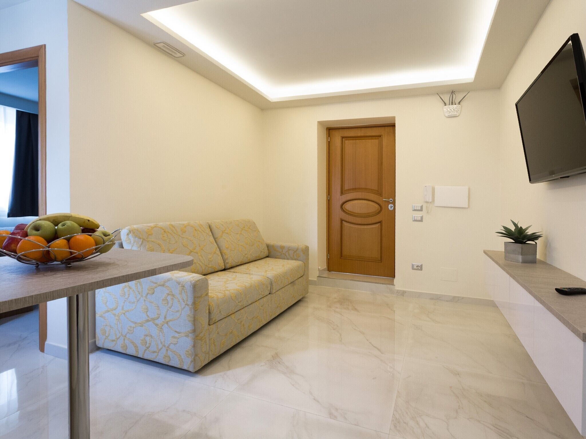 Una sala de estar moderna con un sofá, una mesa de comedor con fruta, una puerta de madera y un televisor de pantalla plana montado en la pared.
