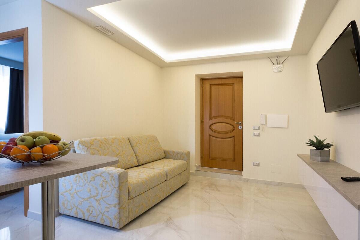 A modern living room with a sofa, a dining table with fruit, a wooden door, and a flat-screen TV mounted on the wall.