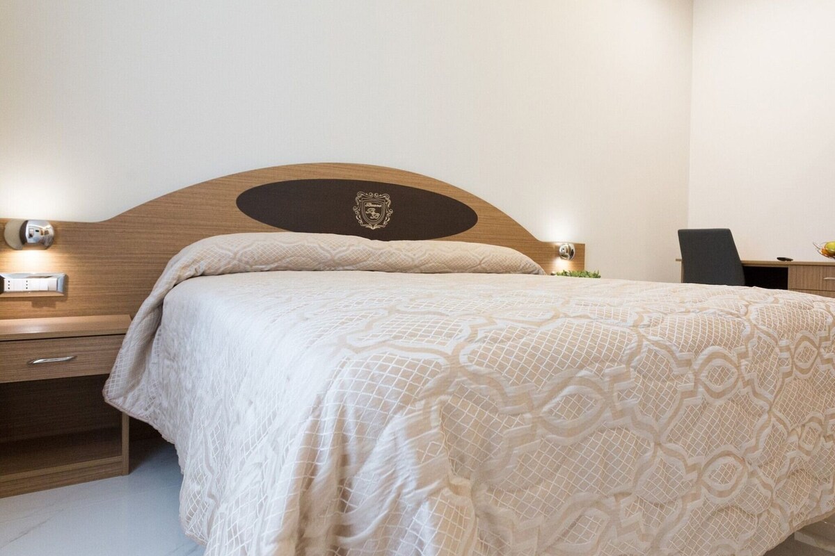 A hotel room with a wooden headboard, a bed with a patterned bedspread, a nightstand with a lamp, and a desk with a chair.