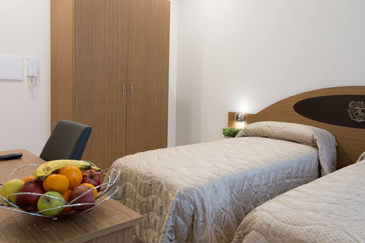 A hotel room with two beds, a wooden wardrobe, a desk with a fruit bowl, and a small bedside table with a lamp.