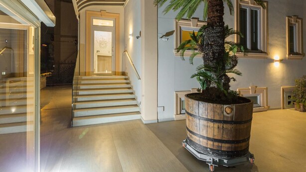 A modern hotel lobby with a palm tree in a barrel planter, a staircase with a curved railing, and a door with a coat of arms. - Buono BB (Naples)