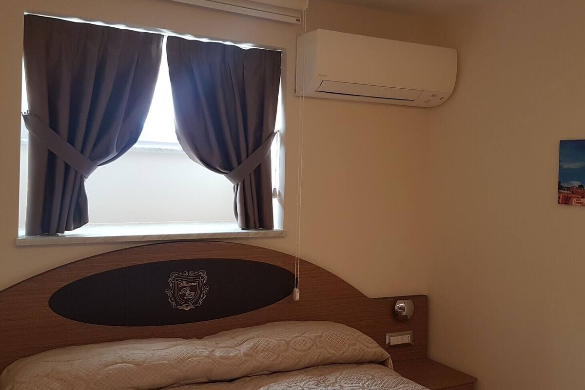 A bedroom with a bed, a window with curtains, an air conditioner, and a wall-mounted light switch.