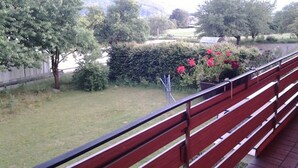 Blick von einem Balkon mit roter Geländer auf eine Rasenfläche mit Bäumen und einer Hecke.