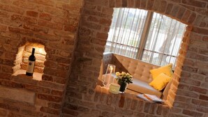 A cozy room with a brick wall, a window with sheer curtains, a sofa with yellow cushions, a bottle of wine, and a candle on a small table. - Do Ciacole in Relais (Mira)