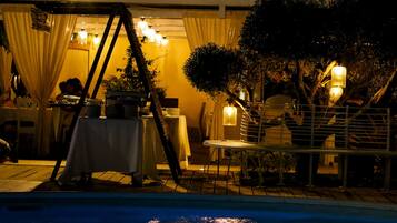 Un área de comedor al aire libre con mesas y sillas, una piscina en primer plano y un edificio con balcón al fondo.