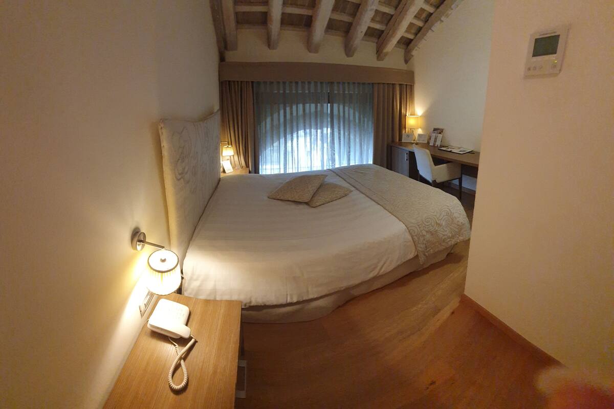 A hotel room with a bed, bedside table with a phone, a desk with a chair, and a window with curtains.