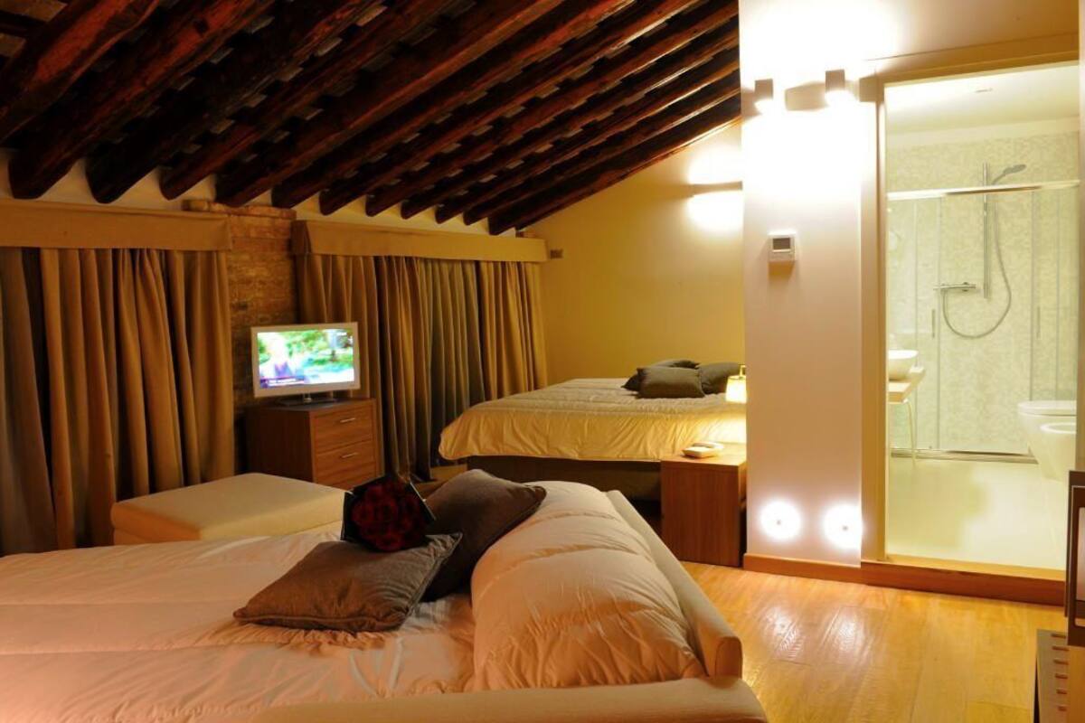 A hotel room with a wooden ceiling, a TV on a wooden stand, two beds, a small table, and a glass-enclosed shower area.