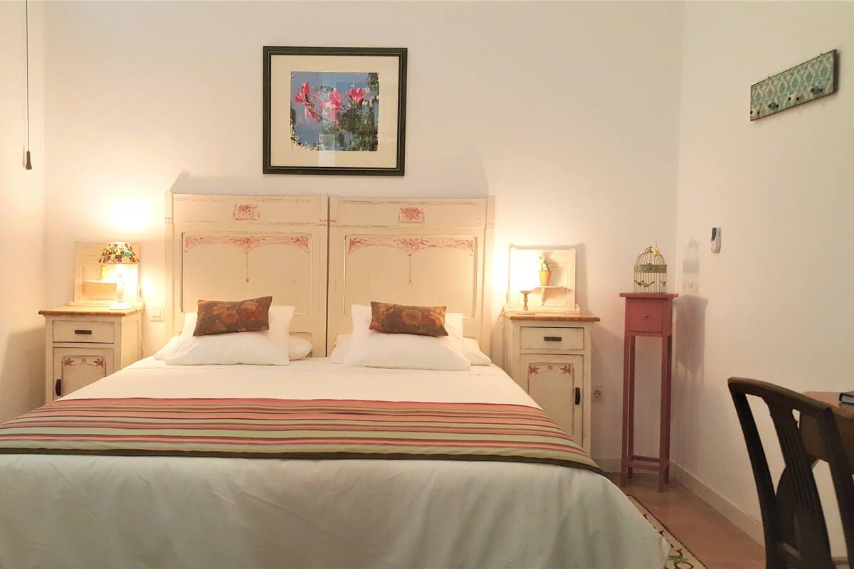 Ein Doppelbett mit gestreifter Bettdecke, zwei Nachttische mit Lampen, ein gerahmtes Bild an der Wand und ein kleiner Tisch mit Vogelkäfig.