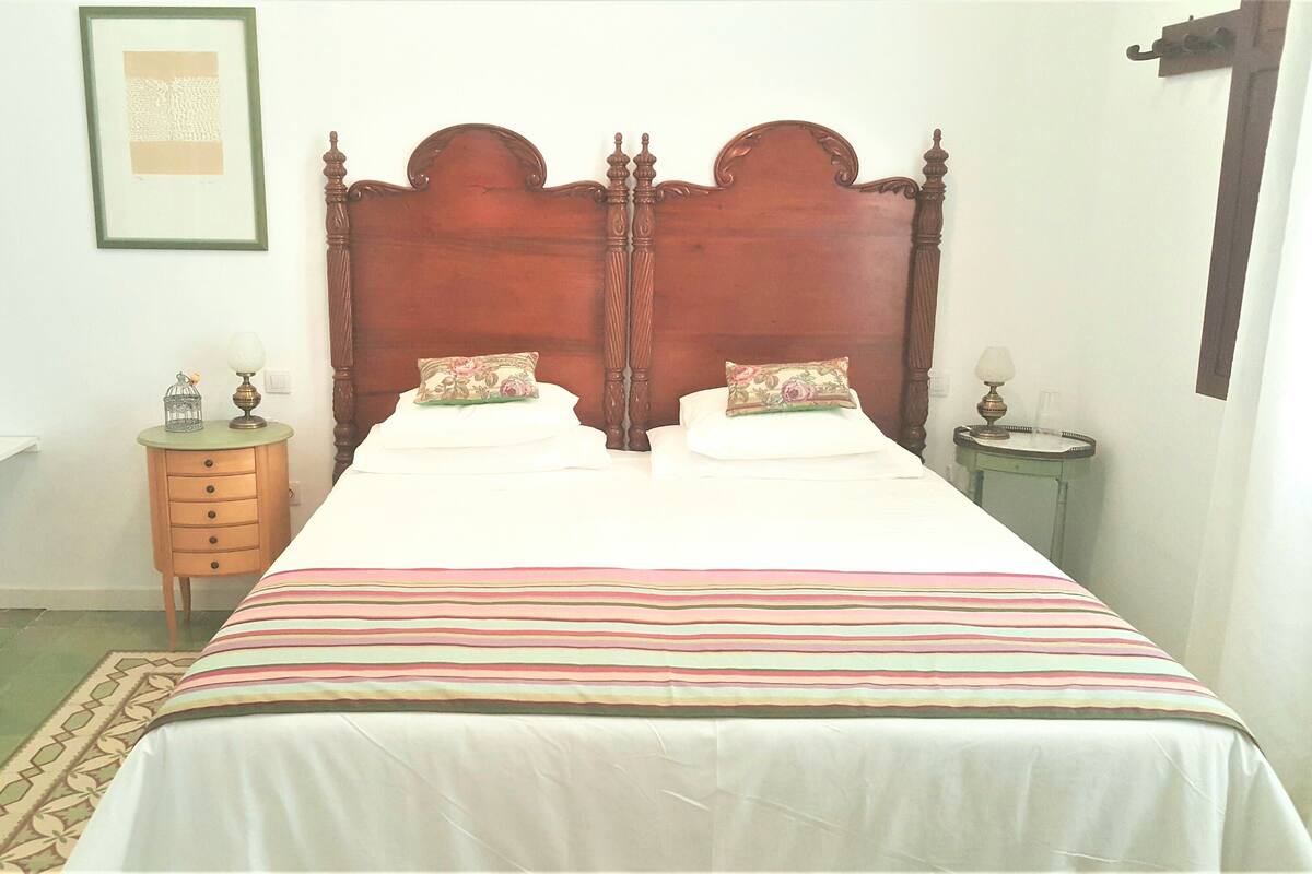Ein Doppelbett mit Holz Kopfteil, zwei Nachttische, ein Beistelltisch, ein gerahmtes Bild an der Wand und ein gemustertes Teppich.