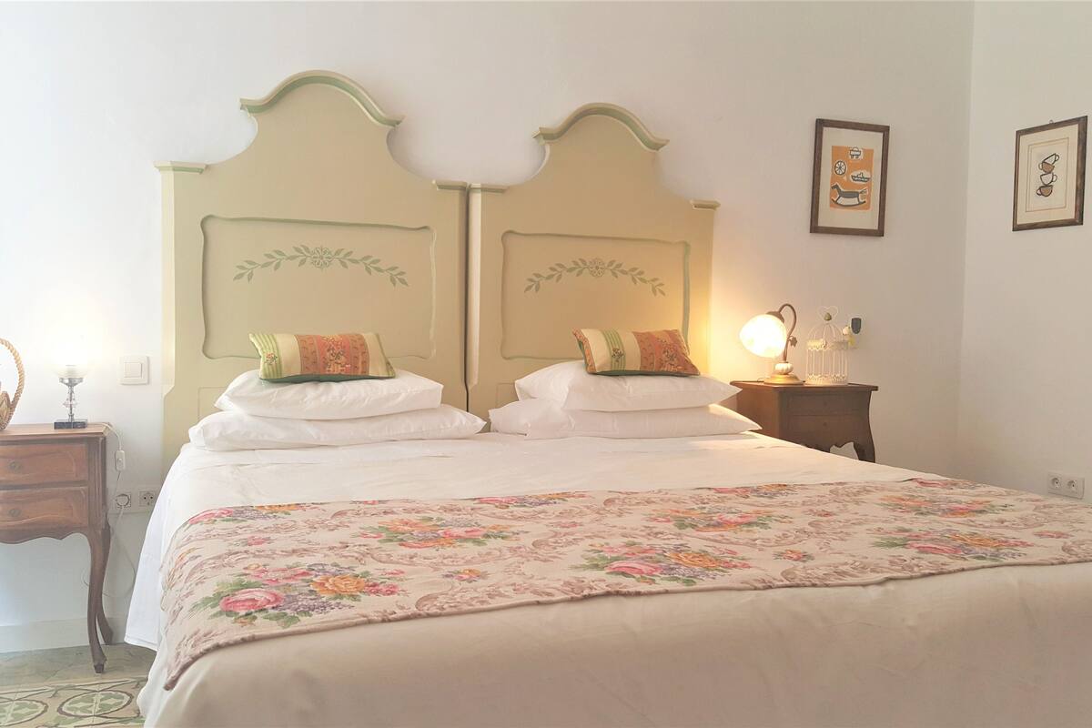 Ein Schlafzimmer mit einem Doppelbett, floralem Bettbezug, zwei Nachttischen, einer Lampe, einer Wanduhr und gerahmten Bildern.