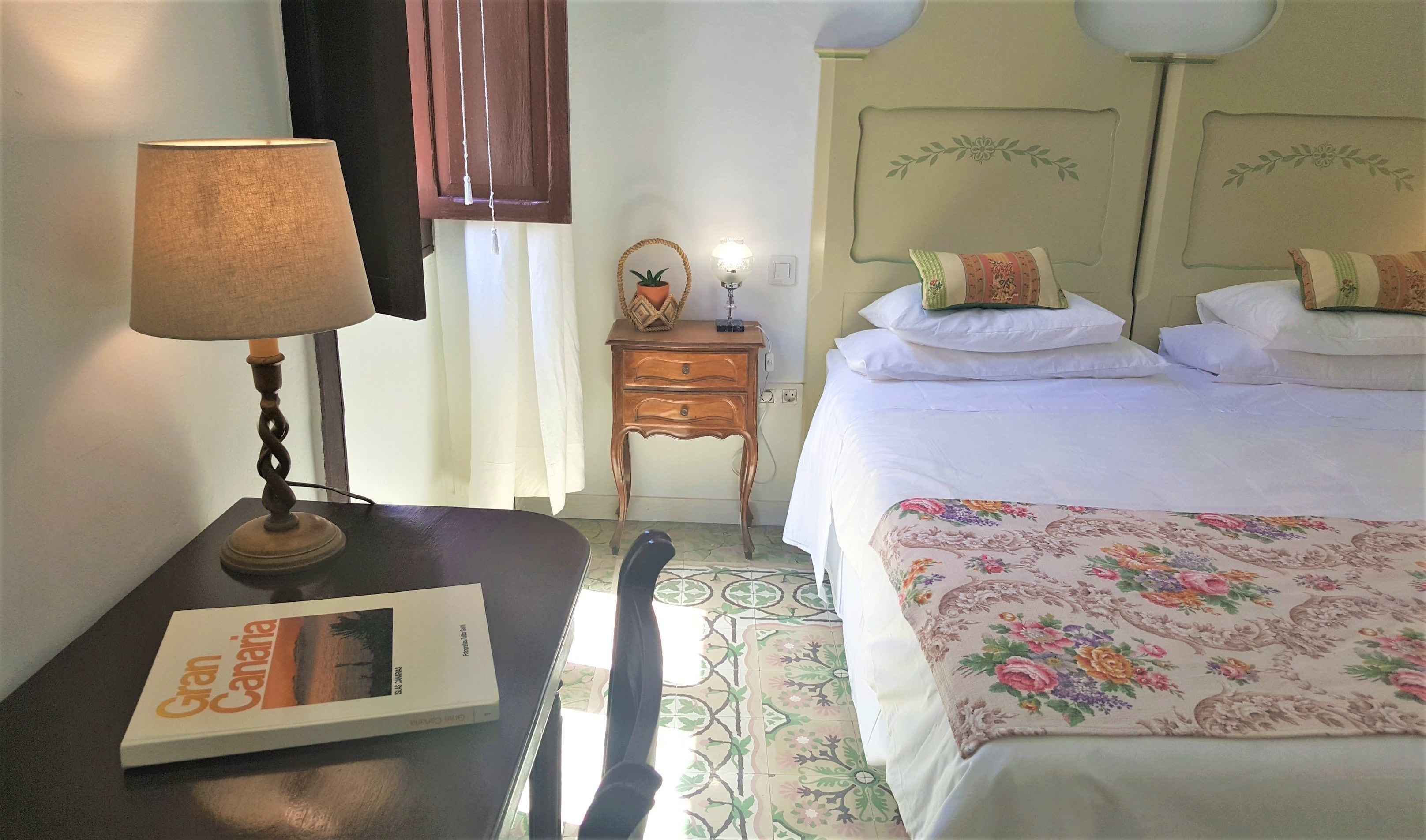 Una habitación de hotel con una cama, una mesita de noche con lámpara, una silla, una pequeña mesita auxiliar de madera y una colcha con estampado floral.