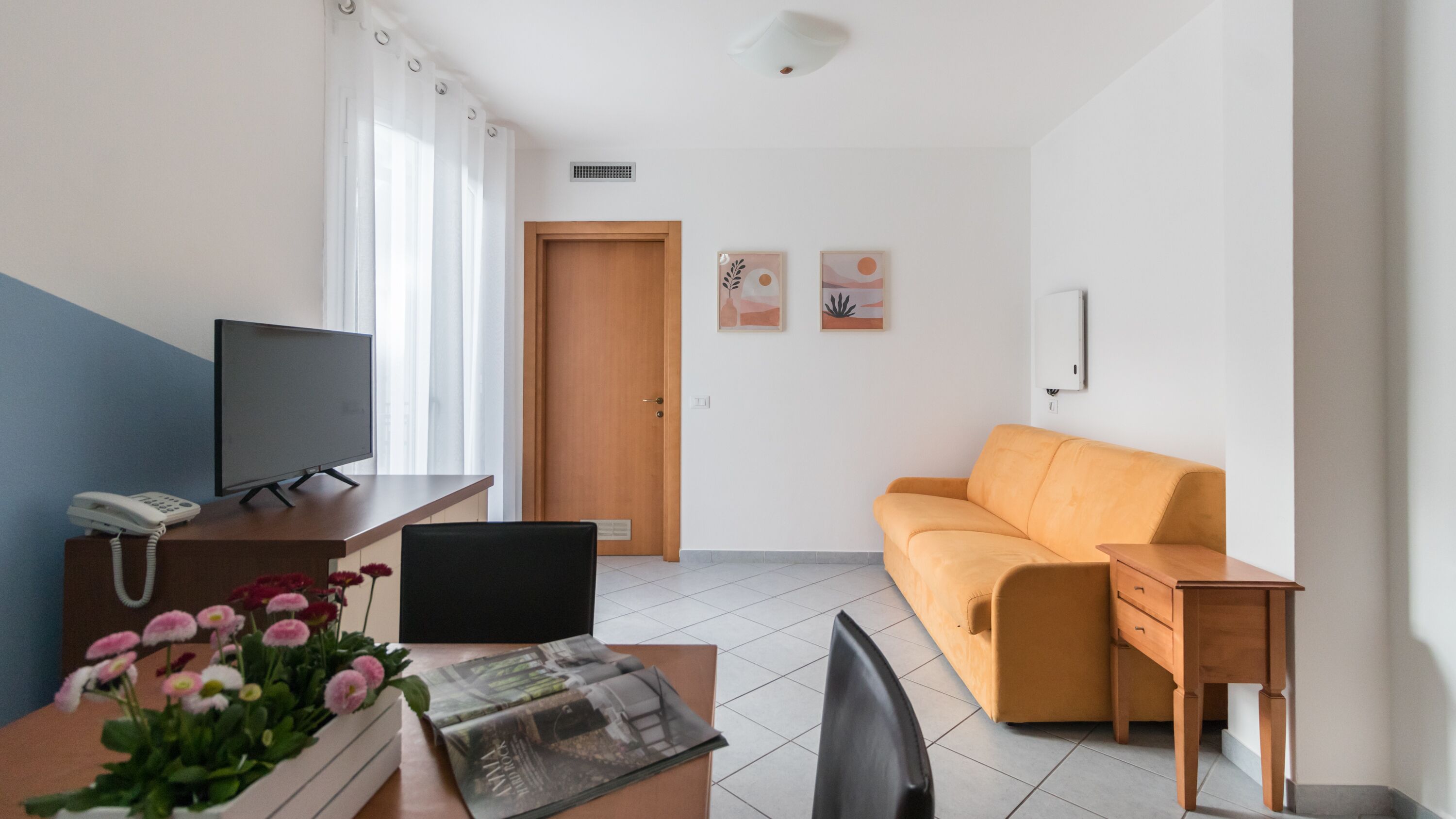 Ein Wohnzimmer mit einer Couch, einem Fernseher, einem Schreibtisch mit Telefon und einem kleinen Tisch mit Blumen.