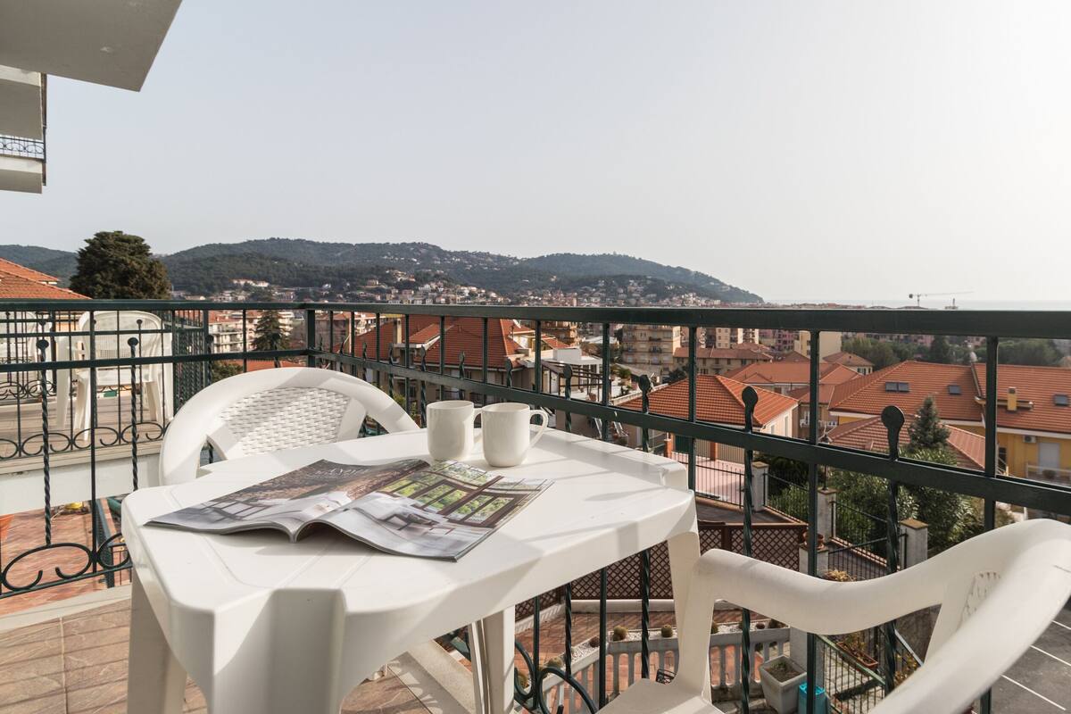 Un balcón con una mesa, sillas y un periódico. El balcón tiene vista a un paisaje urbano con edificios y colinas.