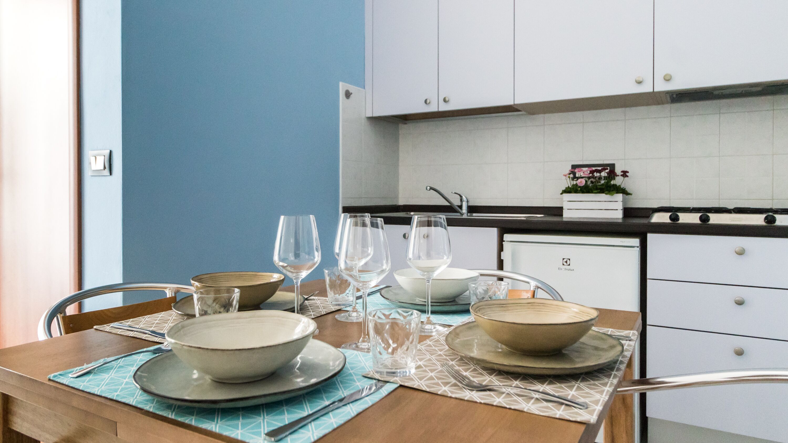 Une table à manger dressée avec des assiettes, des bols et des verres à vin, avec une cuisine en arrière-plan.