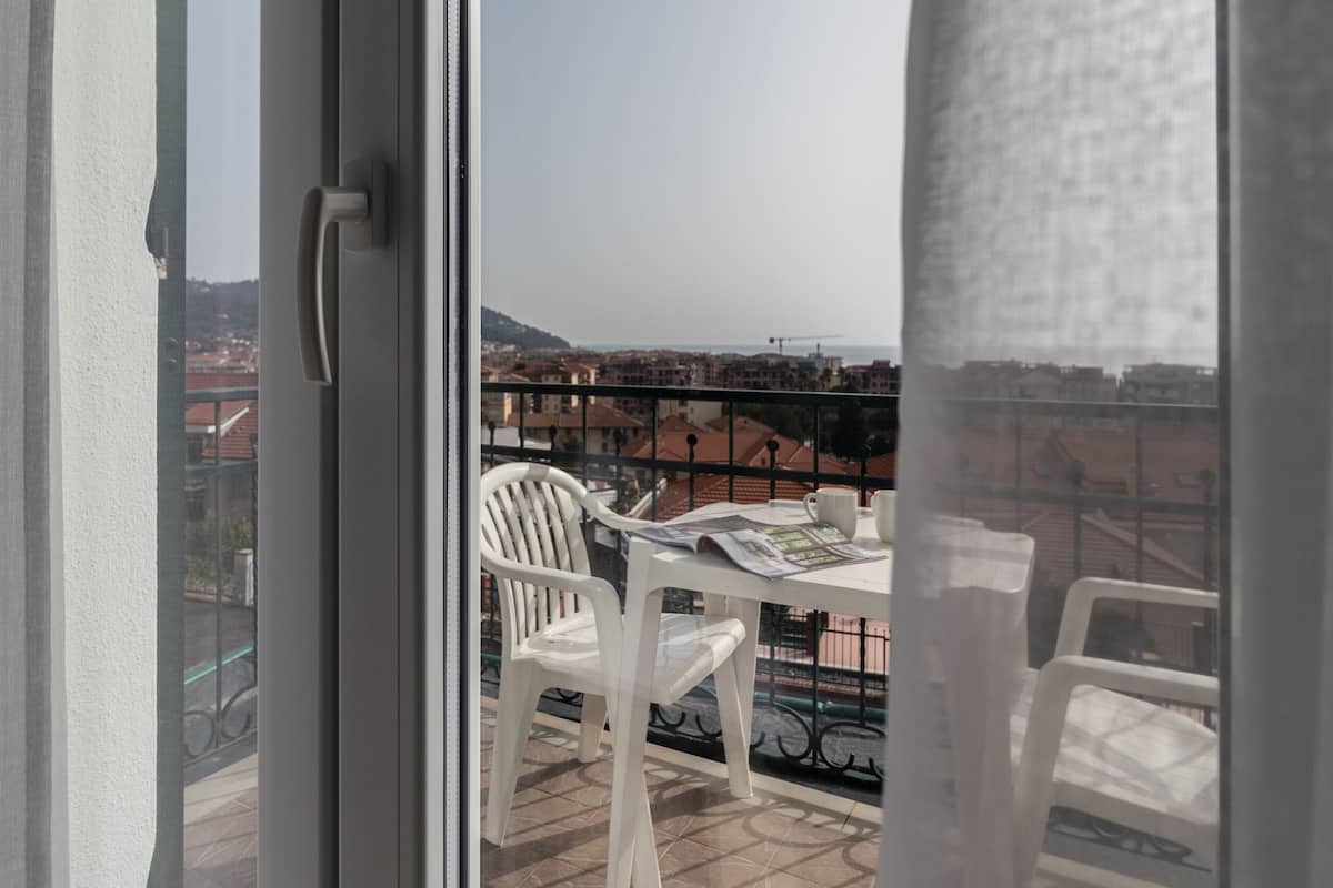 Un balcón con sillas y una mesa de plástico blanco, con vistas a un paisaje urbano con edificios y una vista distante del mar.