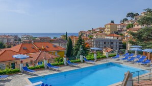 A swimming pool with lounge chairs and umbrellas, surrounded by buildings with red-tiled roofs and a view of the sea. - Residence Il Poggio (Andora)