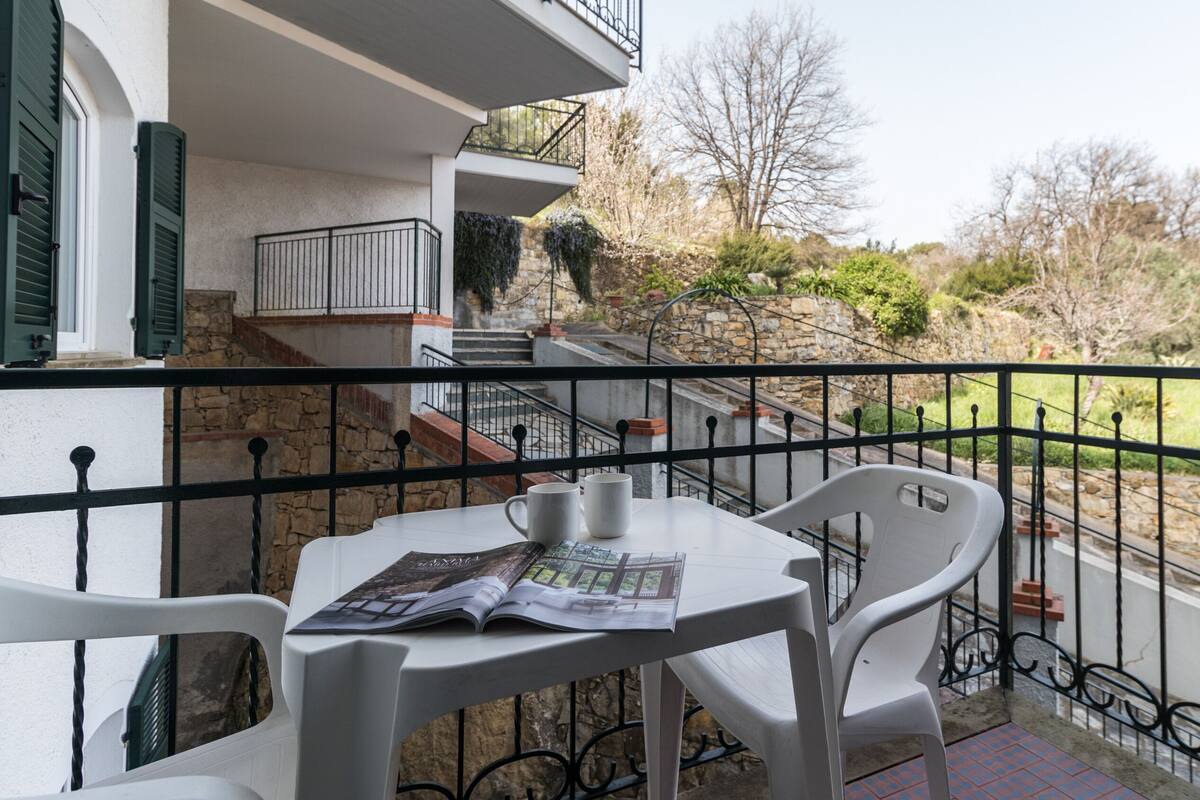 Un balcón con una mesa, sillas, una revista y vistas a un muro de piedra y follaje.