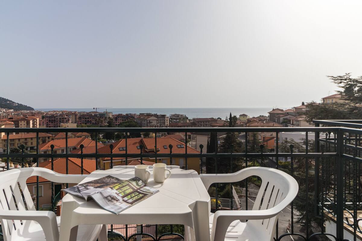 Un balcón con sillas y una mesa de plástico blanco, con vistas a un paisaje urbano costero con edificios de techo rojo y vista al mar.