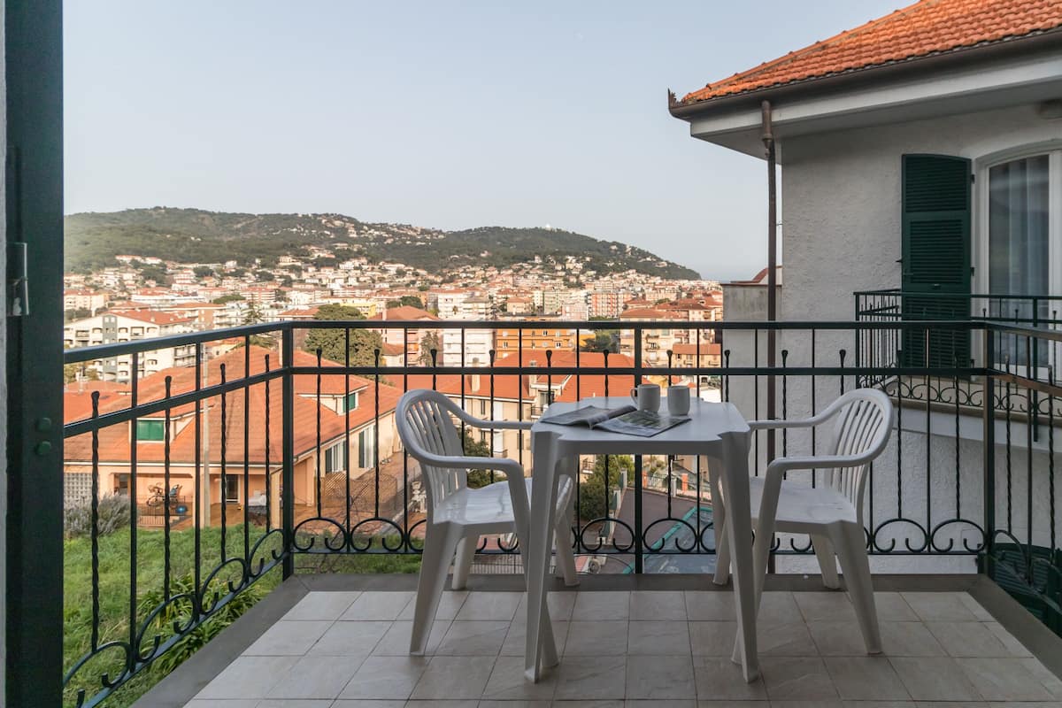 Un balcón con mesa y sillas, con vista a un paisaje urbano que incluye edificios y una colina al fondo.