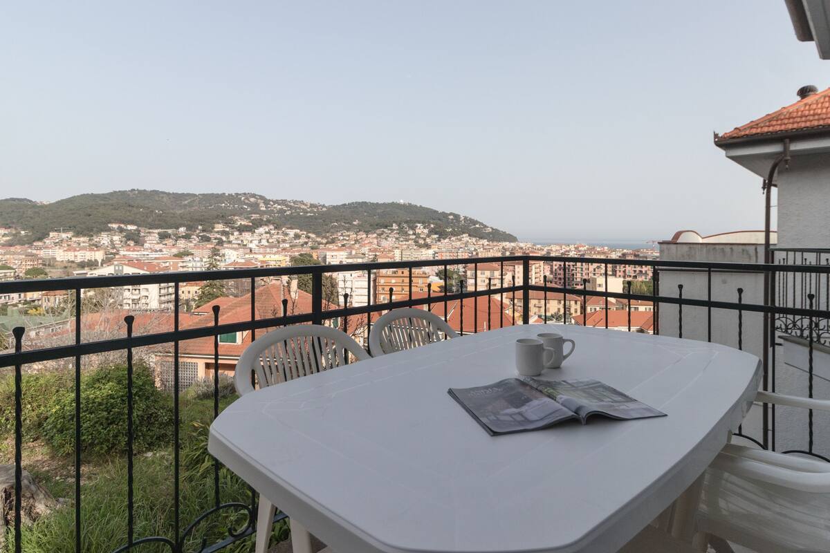 Un balcón con mesa y sillas, con vistas a un paisaje urbano que incluye edificios y colinas.