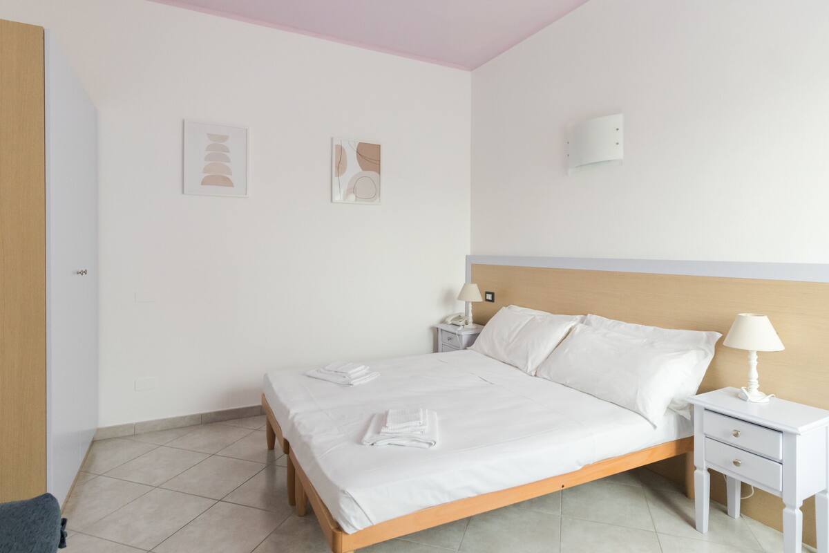 Una cama bien hecha con sábanas blancas, cabecera de madera, mesita de noche con lámpara y dos apliques de pared.