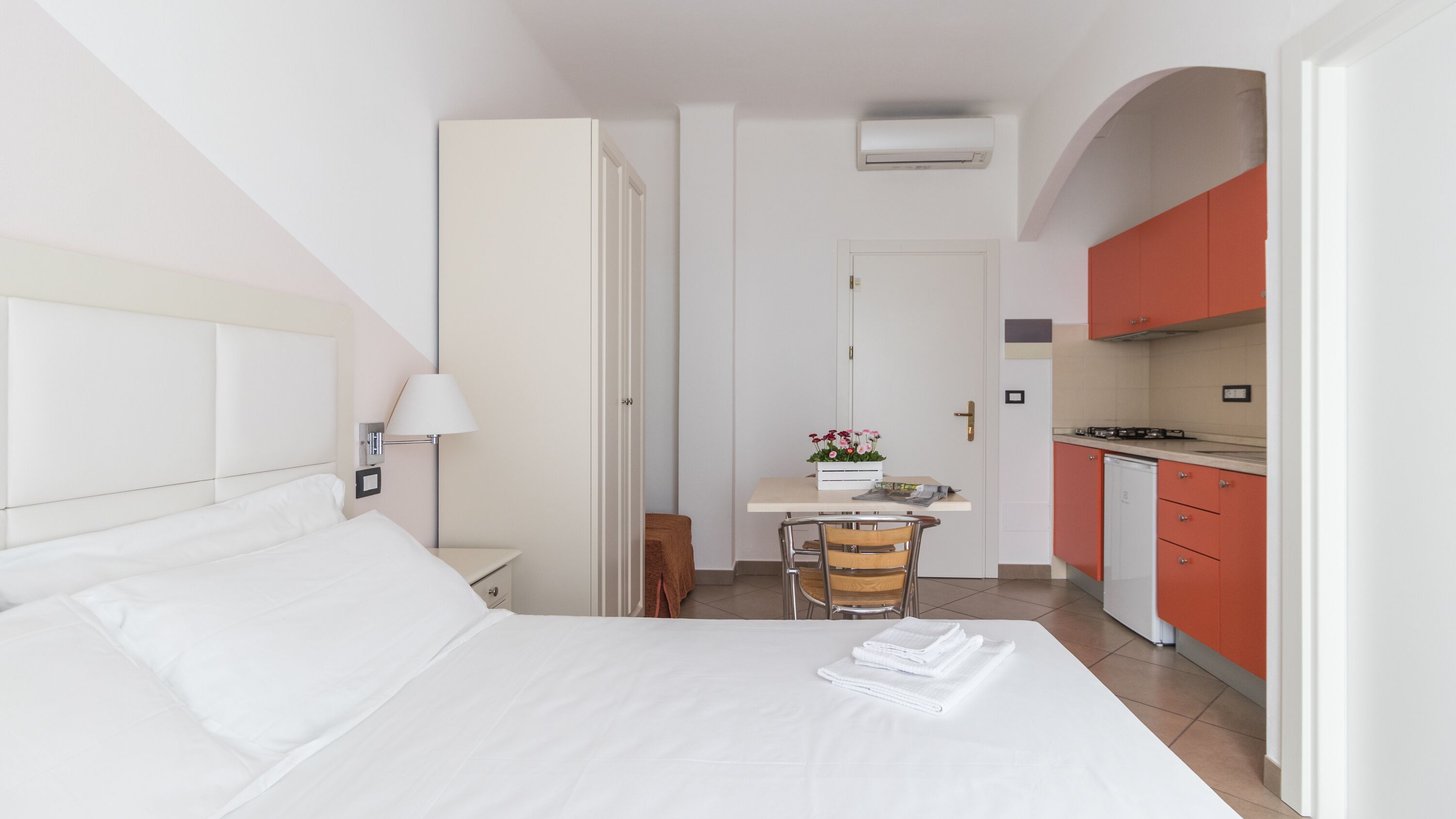 Ein Hotelzimmer mit einem Bett, einem Nachttisch mit Lampe, einem kleinen Schreibtisch mit Stuhl und einer Küchenzeile mit roten Schränken und einem Herd.