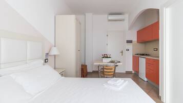 Ein Hotelzimmer mit einem Bett, einem Nachttisch mit Lampe, einem kleinen Schreibtisch mit Stuhl und einer Küchenzeile mit roten Schränken und einem Herd.