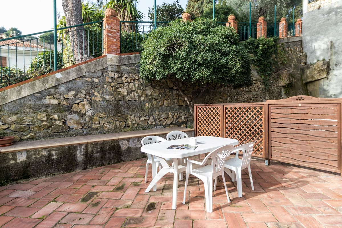 Un patio con muro de piedra, cerca de madera y mesa blanca con sillas.