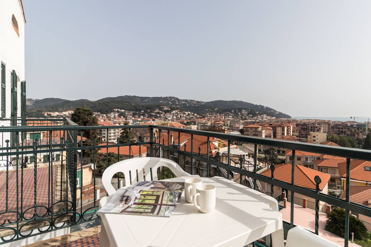Un balcón con mesa y sillas, con vista a un pueblo costero de edificios con tejados de tejas rojas y vistas al mar.