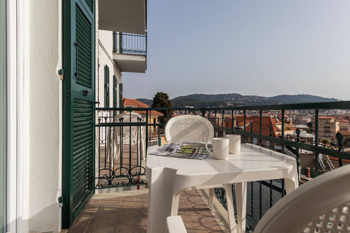Un balcón con mesa y sillas, con vista a un paisaje urbano que incluye edificios y colinas.