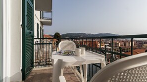 Un balcón con mesa y sillas, con vista a un paisaje urbano que incluye edificios y colinas.