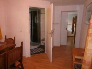 A room with a wooden dining table and chairs, a shower area, and a hallway leading to another room.
