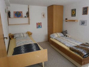 A small, compact room with two single beds, a wooden wardrobe, and a shelf with various items.