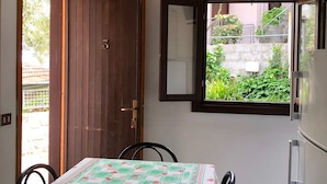 A room with a wooden door, an air conditioning unit, a window with a view of greenery, a table with a patterned tablecloth, and a refrigerator. - Villaggio Turistico Dea Cupra (Cupra Marittima)