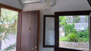 A room with a wooden door marked with the number 3, a large window with a view of greenery, and a wall-mounted air conditioning unit. - Villaggio Turistico Dea Cupra (Cupra Marittima)