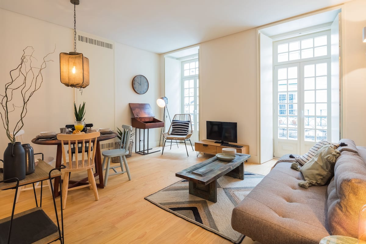 Un salon moderne avec un canapé, une table à manger et des chaises. On y trouve également un téléviseur, une horloge et une fenêtre donnant sur des immeubles.