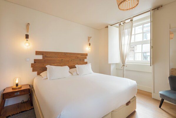 A bedroom with a wooden headboard, a large bed with white linens, two bedside lamps, and a window with curtains.