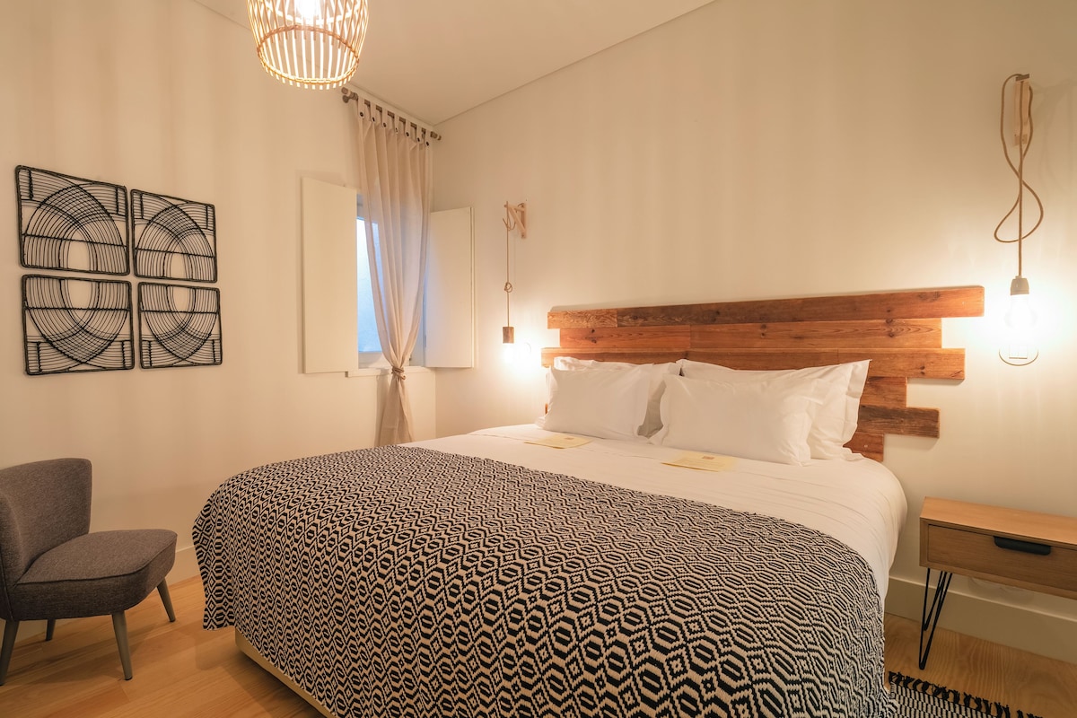 Quarto bem arrumado com colcha estampada, cabeceira de madeira, mesa de cabeceira e uma cadeira.