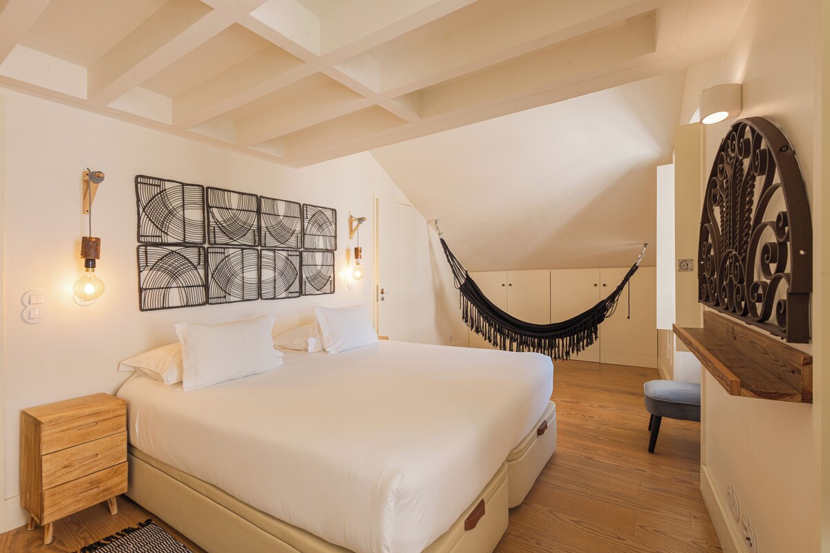 A bedroom with a large bed, a wooden nightstand, a wall-mounted light fixture, and a hammock.