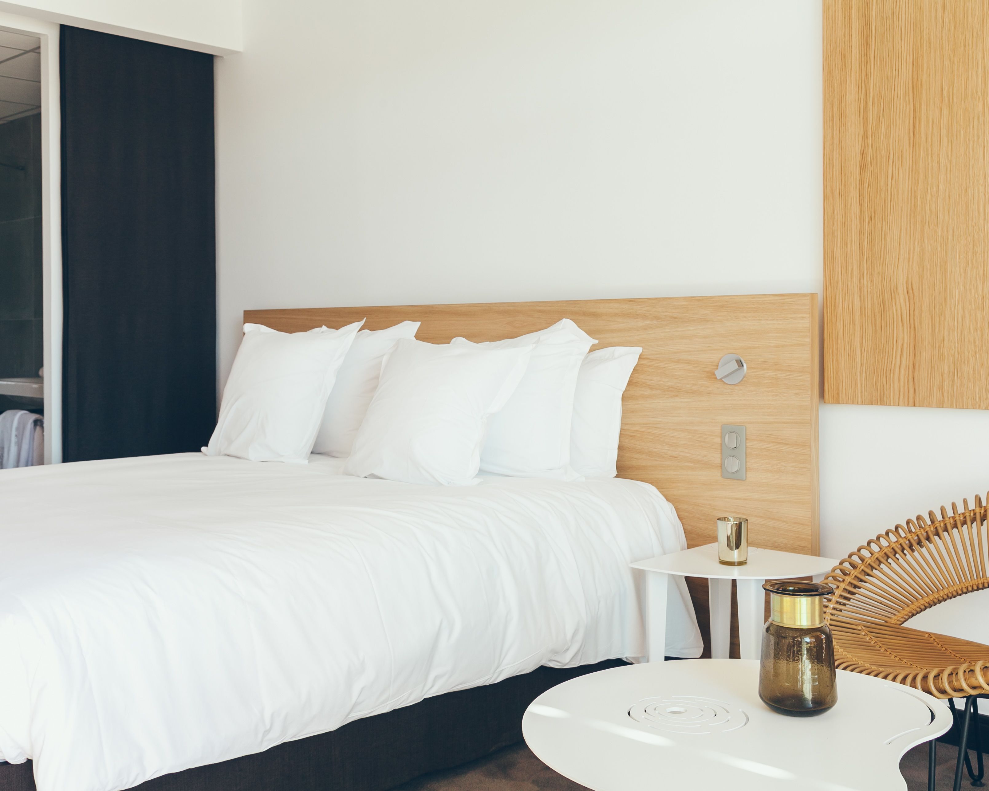 Una habitación de hotel moderna con una cama grande, ropa de cama blanca, una mesita de noche pequeña con una lámpara y una silla de mimbre.