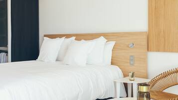 Una habitación de hotel moderna con una cama grande, ropa de cama blanca, una mesita de noche pequeña con una lámpara y una silla de mimbre.