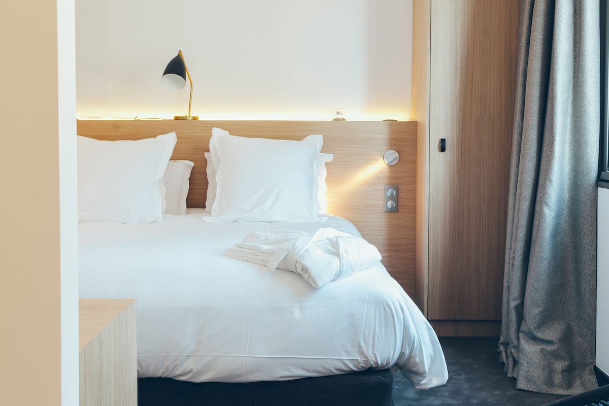 A hotel room with a neatly made bed, a bedside lamp, a wooden headboard, and a door with curtains.