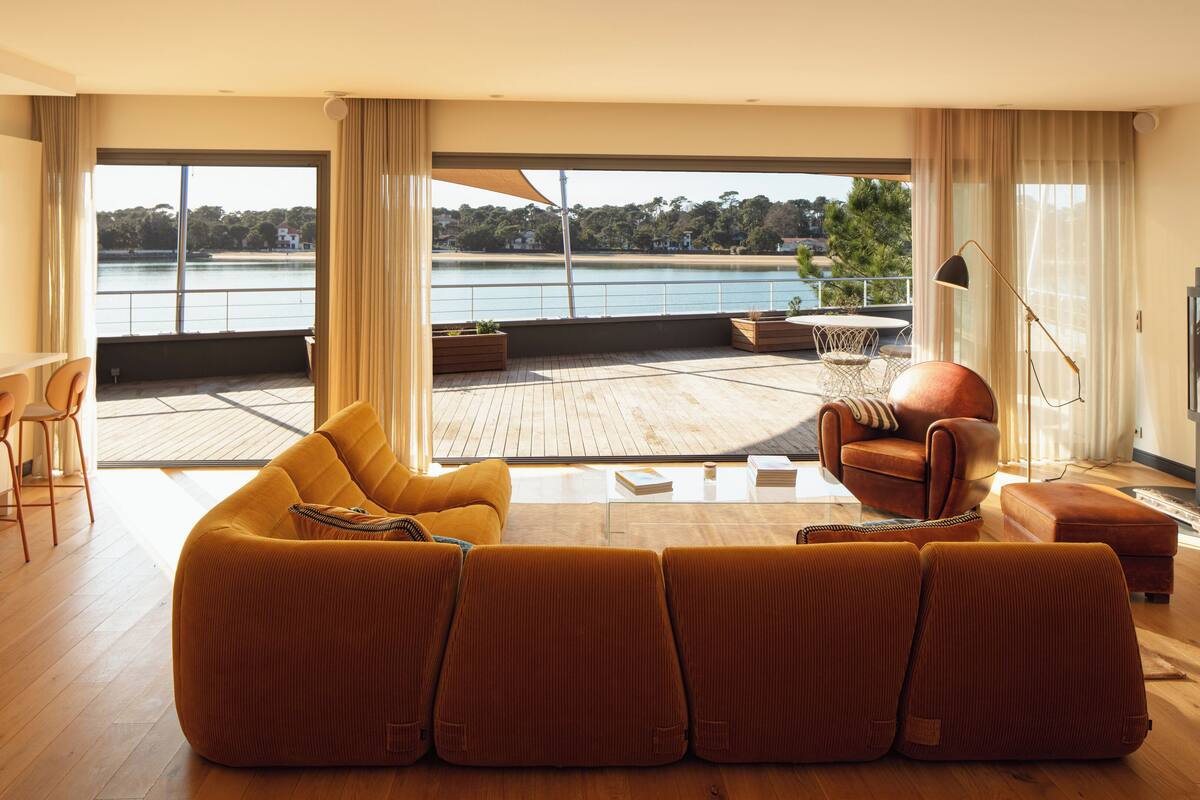 A spacious living room with a large sofa, a chair, and a view of a lake through large windows.