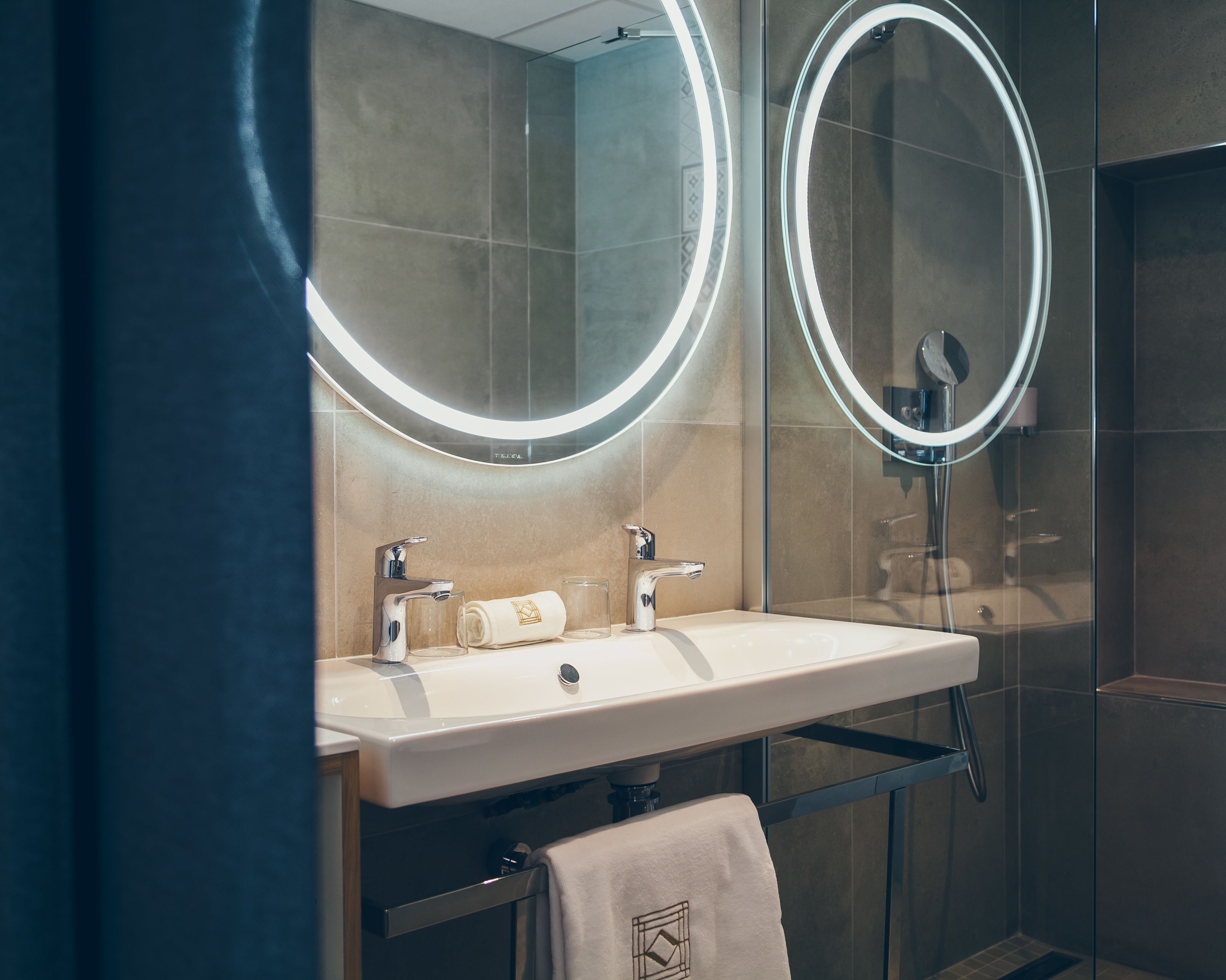 A modern bathroom with a white sink, two mirrors with illuminated frames, a towel rack, and a glass shower enclosure.