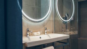 A modern bathroom with a white sink, two mirrors with illuminated frames, a towel rack, and a glass shower enclosure.