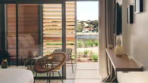 Una habitación de hotel moderna con un amplio ventanal corredero que da a un balcón, una cama, una silla de mimbre, una mesita con un florero y un televisor fijado a la pared.