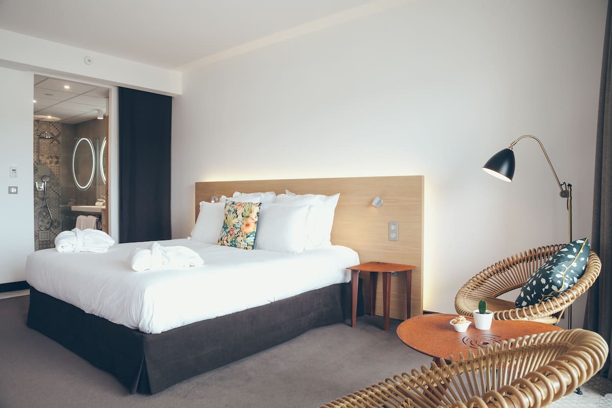 A modern hotel room with a neatly made bed, a wicker chair, a bedside table with a lamp, and a view of the bathroom.