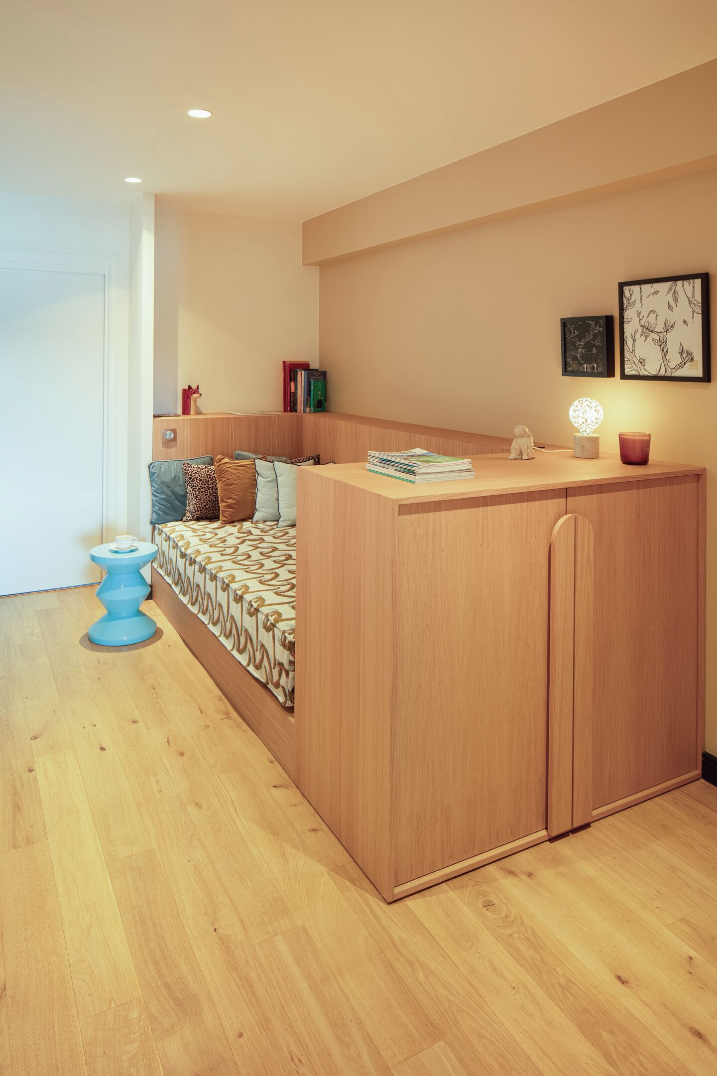 A compact room with a bed, a small desk, a blue stool, and a wall-mounted shelf with books and decorative items.