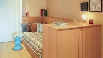 A compact room with a bed, a small desk, a blue stool, and a wall-mounted shelf with books and decorative items.