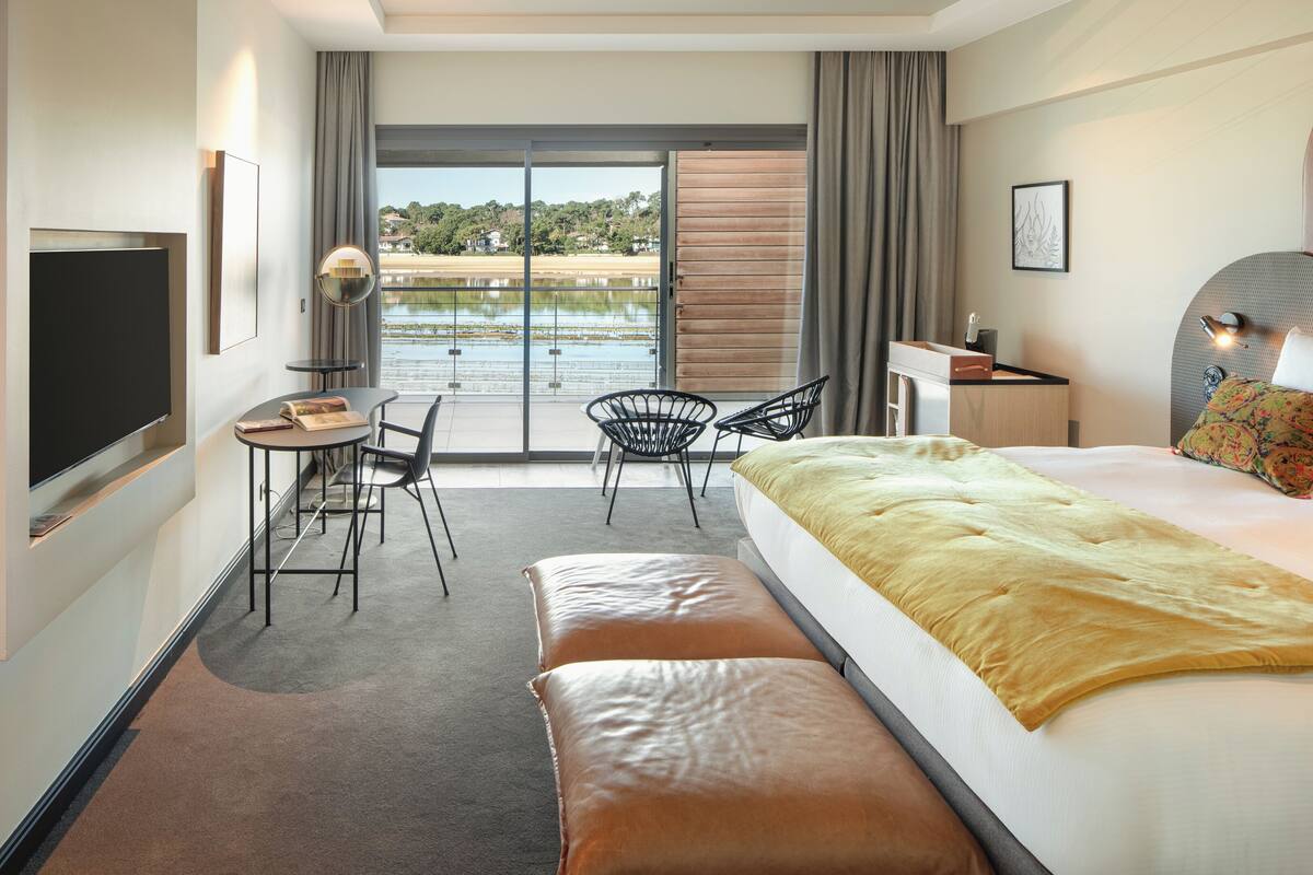 A modern hotel room with a large bed, a desk, a chair, and a view of a body of water through a sliding door.